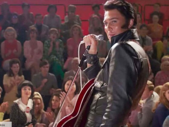 Elvis is looking back at the camera with a smile, microphone in hand, dressed in the same black leather jacket as the movie poster with red guitar in hand.